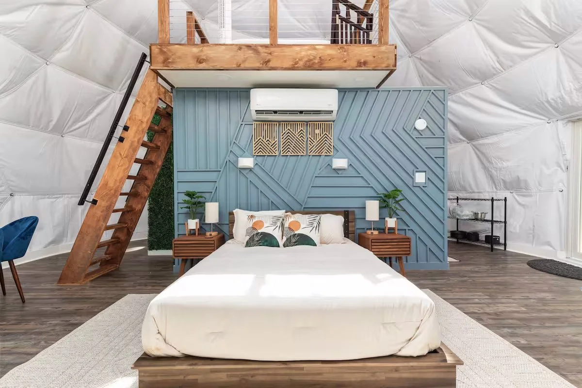 Modern bedroom with a wooden staircase, a blue decorative wall behind the bed, and a loft area above. The bed has white sheets and decorative pillows, with matching nightstands and lamps. There are potted plants on the nightstands and a small rack to the right.