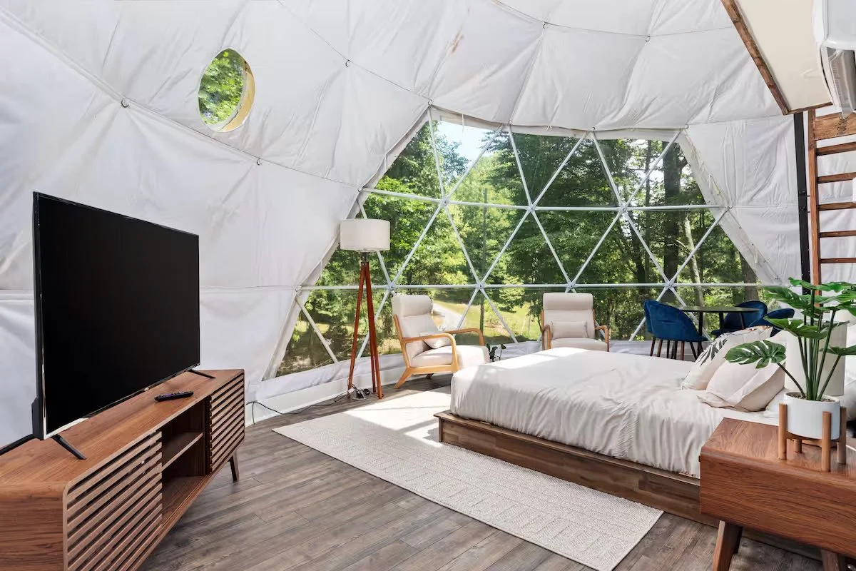 Interior of a geodesic dome bedroom with large glass windows, a bed, a TV on a wooden stand, a side table with plants, two armchairs, and some chairs near the window, with greenery visible outside.