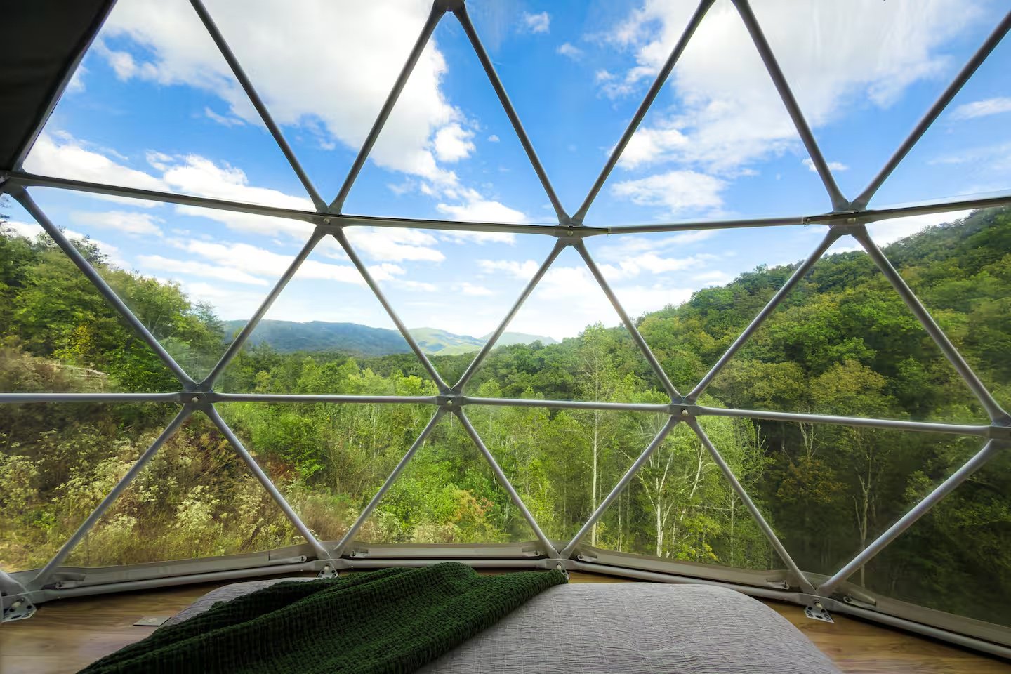 View from inside a geodesic dome overlooking a lush green forest and rolling hills under a partly cloudy sky.