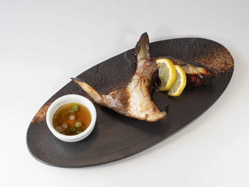 Grilled fish served with lemon slices, garnished with red chili threads, accompanied by a small bowl of dipping sauce with green onions on a black oval plate.