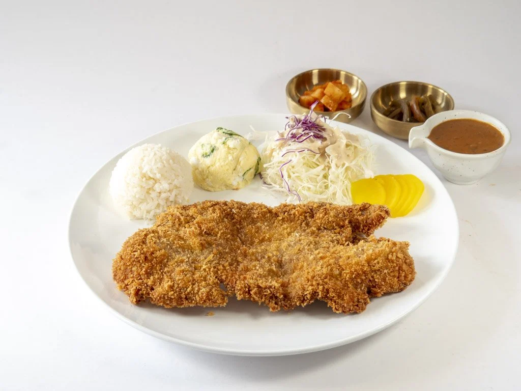 Plate with fried fish, rice, potato salad, shredded cabbage, and pickles, with side bowls of kimchi, pickled vegetables, and dipping sauce on white background.