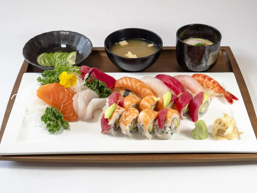Sushi platter with various nigiri and rolls, accompanied by wasabi, pickled ginger, cucumber slices, and three bowls of miso soup on a wooden tray.