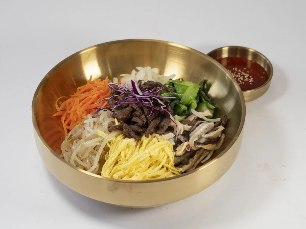 A metallic bowl filled with assorted vegetables, beef, and noodles, accompanied by a small bowl of red sauce with sesame seeds.