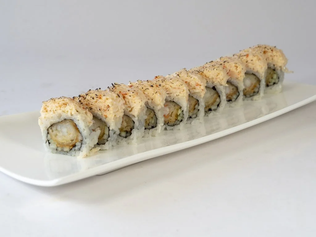A row of sushi rolls on a white rectangular plate, with visible rice, seafood, and possibly avocado or cucumber inside, topped with a crunchy coating and seasonings.