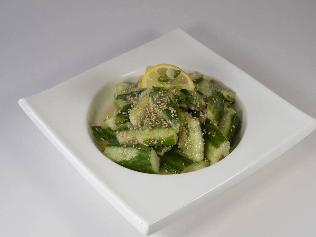 Cucumber salad garnished with sesame seeds and a lemon wedge on a white square plate.