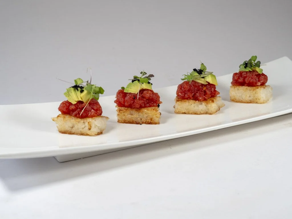 Four bite-sized appetizers on a white rectangular plate, each with a toasted base, topped with tomato-based mixture, a slice of avocado, and microgreens.
