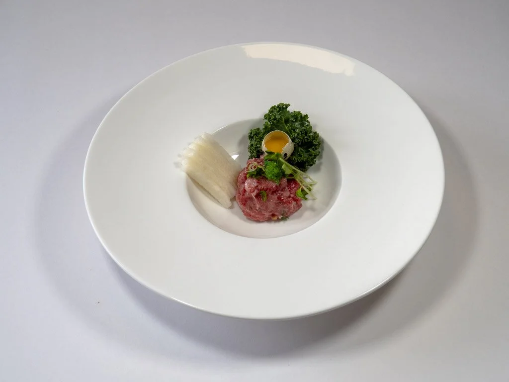 Small portion of raw tuna tartare garnished with greens, a slice of white radish, kale, and a small yellow sauce container, served on a white plate.