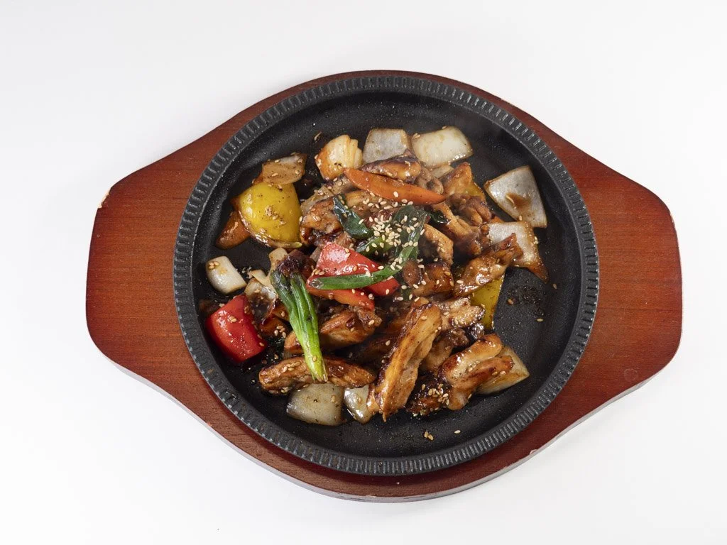 A sizzling skillet with fried chicken and vegetables like peppers and onions, garnished with sesame seeds.
