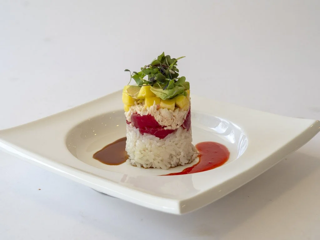 A sushi roll topped with microgreens, served on a white plate with two sauces, one dark brown and one red.