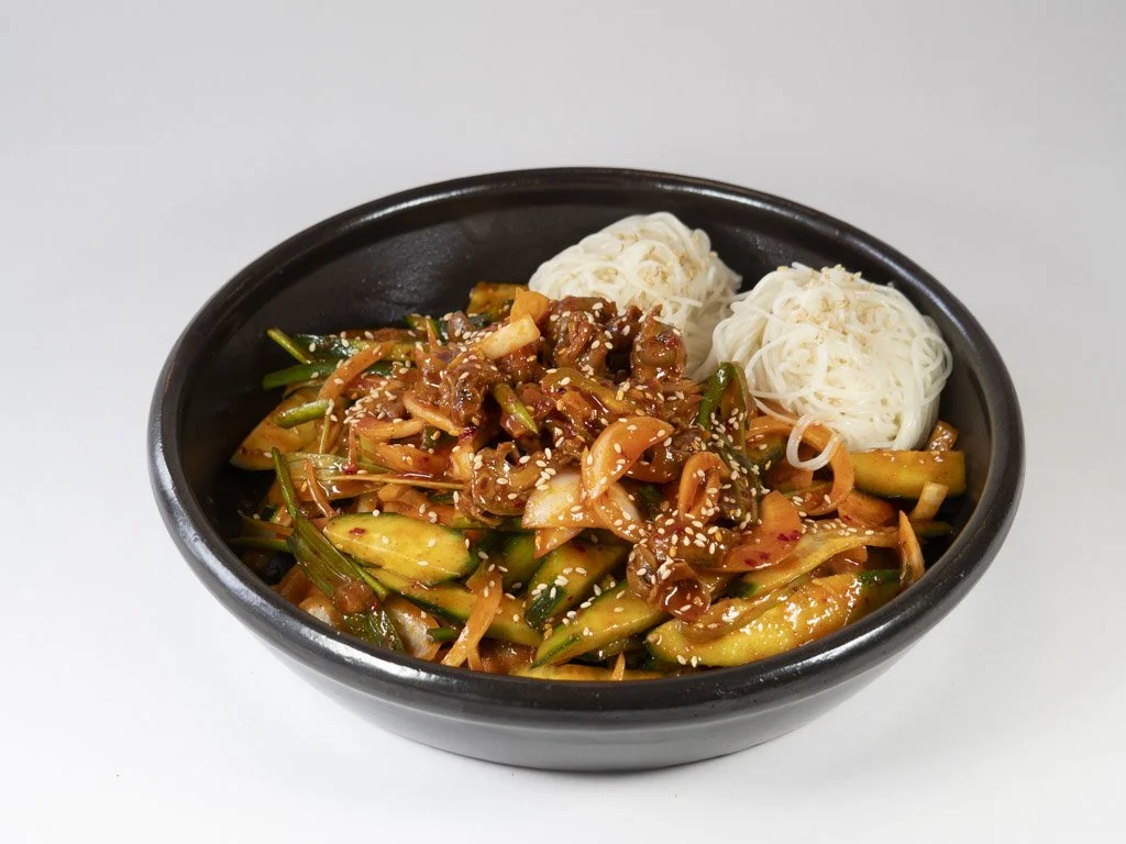 Seafood stir-fry with zucchini, carrots, and green onions, topped with sesame seeds, served with two portions of rice noodles in a black bowl.