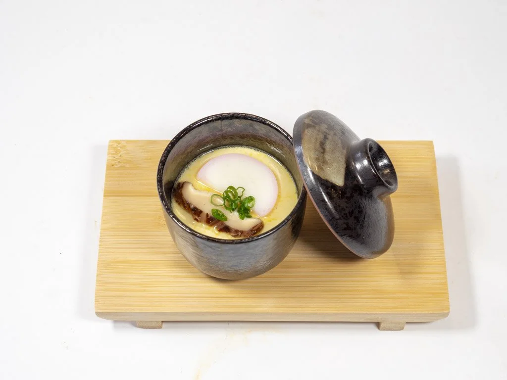 A black pot with a lid slightly open, placed on a wooden tray, containing a Japanese-style chawanmushi with a slice of fish cake, green onions, and other ingredients, on a white surface.