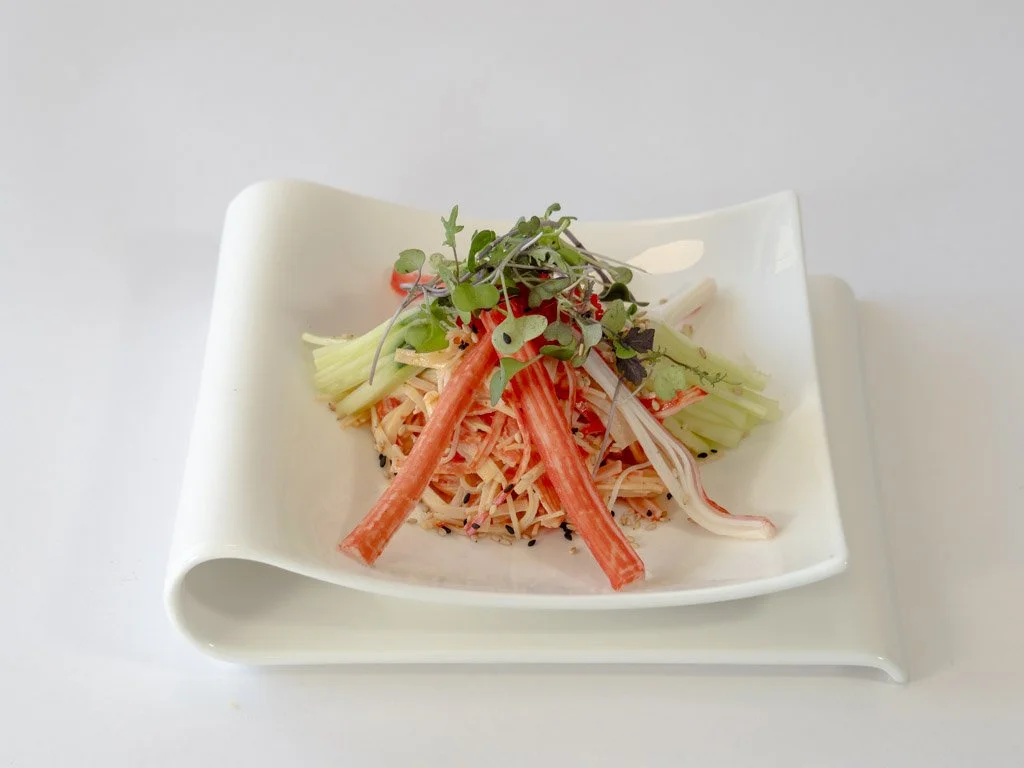 A plate of crab and vegetable noodles garnished with microgreens on a white, curved dish.
