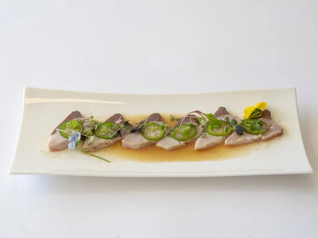 A white rectangular plate with slices of raw fish, garnished with microgreens and edible flowers, served with a light sauce.