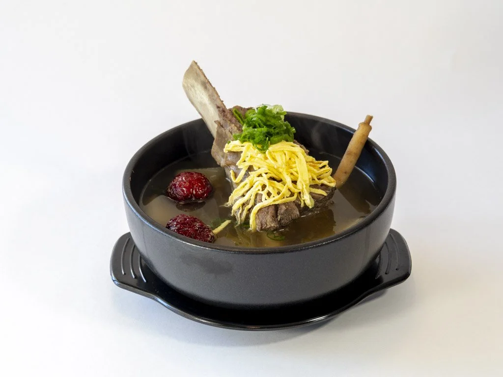 A bowl of clear soup with pieces of shredded yellow cheese, green onions, red berries, and a piece of bone with meat attached.