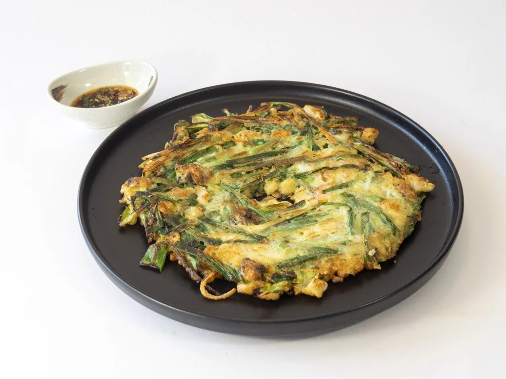 Vegetable pancake with green onions on a black plate, served with soy sauce in a small white bowl.