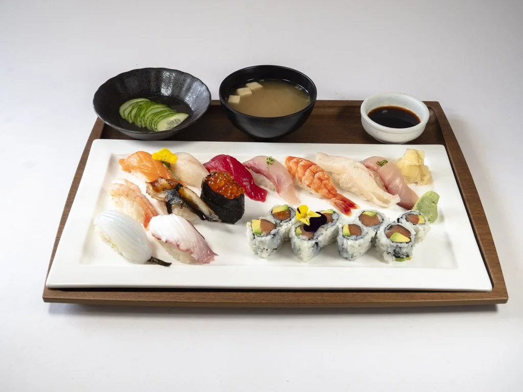 A platter of assorted sushi with sliced cucumbers, miso soup, soy sauce, and wasabi and ginger garnishes on a wooden tray.