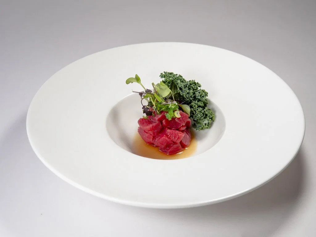 A white plate with a hole in the center containing raw tuna, topped with microgreens, and garnished with kale and a light sauce or broth.