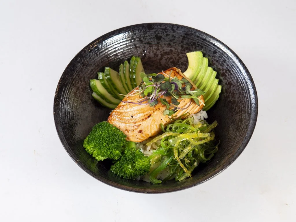 Salmon sashimi bowl with cucumber slices, broccoli, and seaweed, garnished with microgreens, served in a black bowl