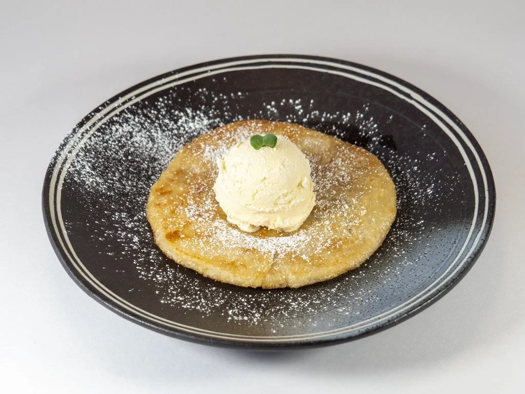 A plate with a cooked apple pancake topped with a scoop of vanilla ice cream, garnished with a small green herb, and dusted with powdered sugar.