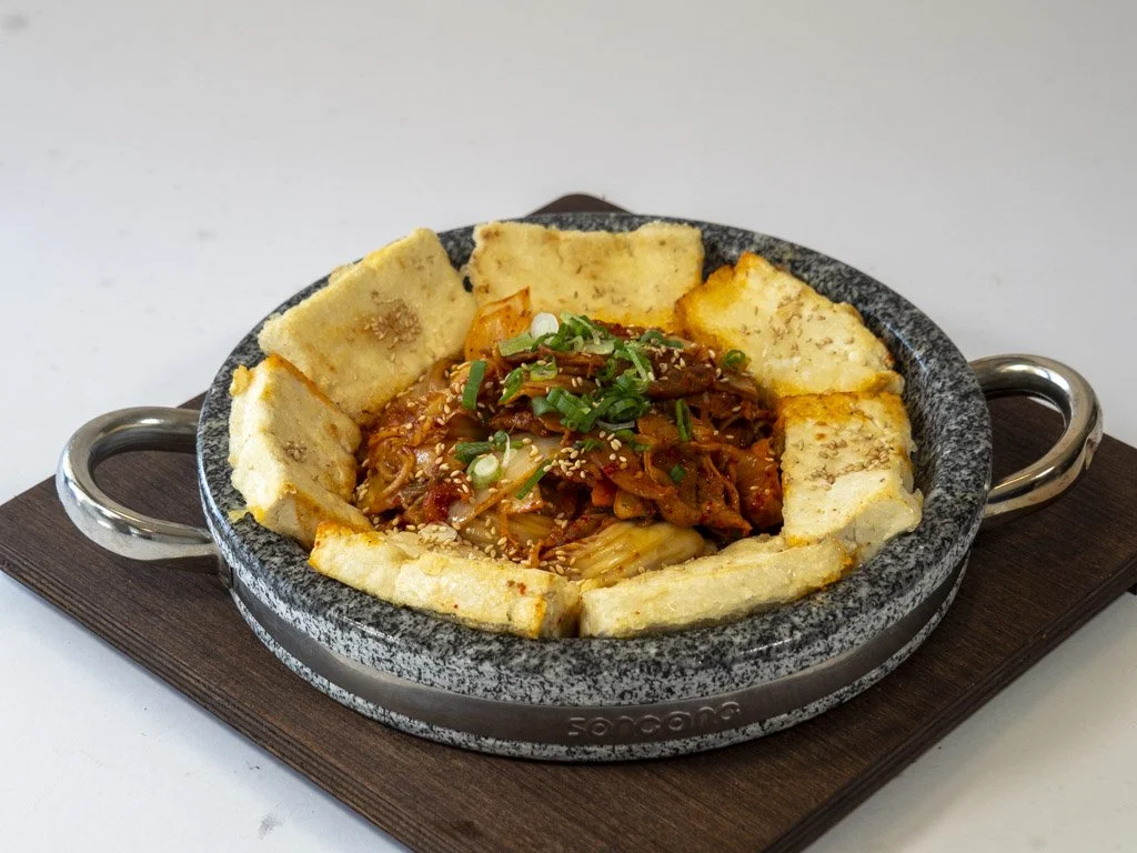 Stone bowl of Korean spicy stew with tofu and vegetables, garnished with green onions and sesame seeds, surrounded by fried tofu pieces.
