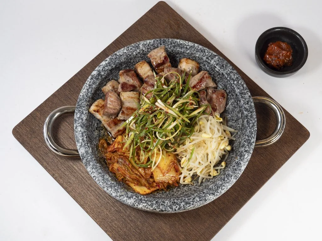 A stone bowl filled with sliced pork belly, kimchi, bean sprouts, and green onion slices, served on a wooden board with a small jar of spicy red sauce.