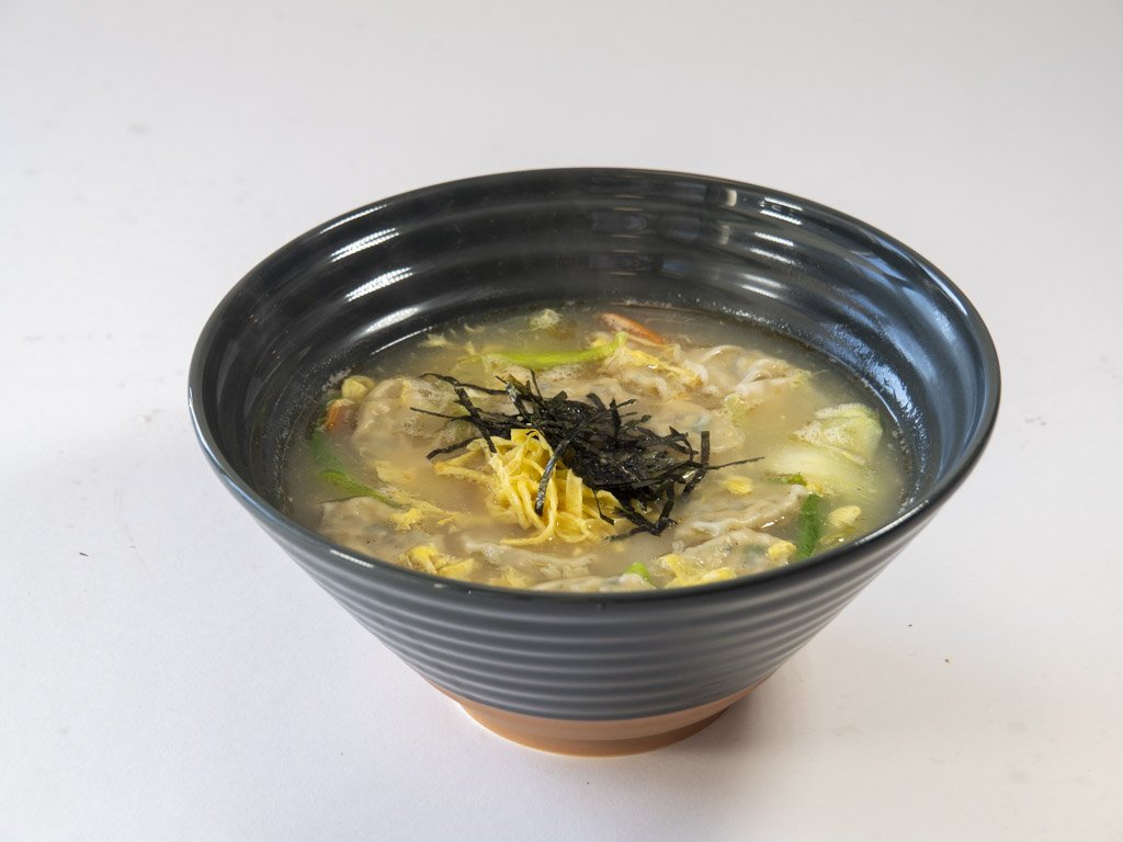 A black bowl of chicken and vegetable soup garnished with shredded nori seaweed and shredded cheese, on a white background.