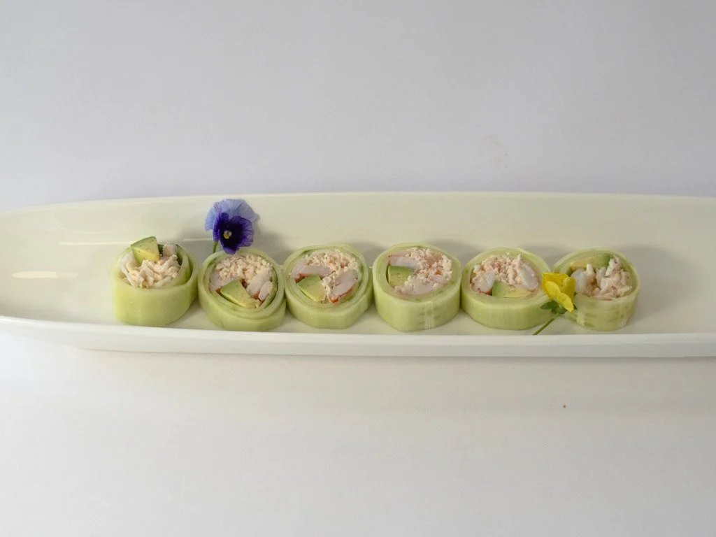 Six cucumber rolls filled with crab salad and garnished with edible flowers on a white oval plate.