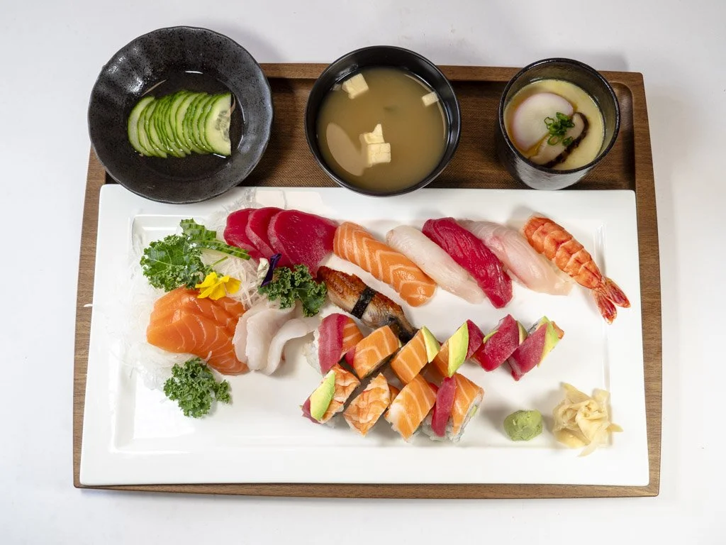 Assorted sushi platter with sashimi, topped with slices of avocado, served with cucumber, miso soup, and dipping sauces on a wooden tray.