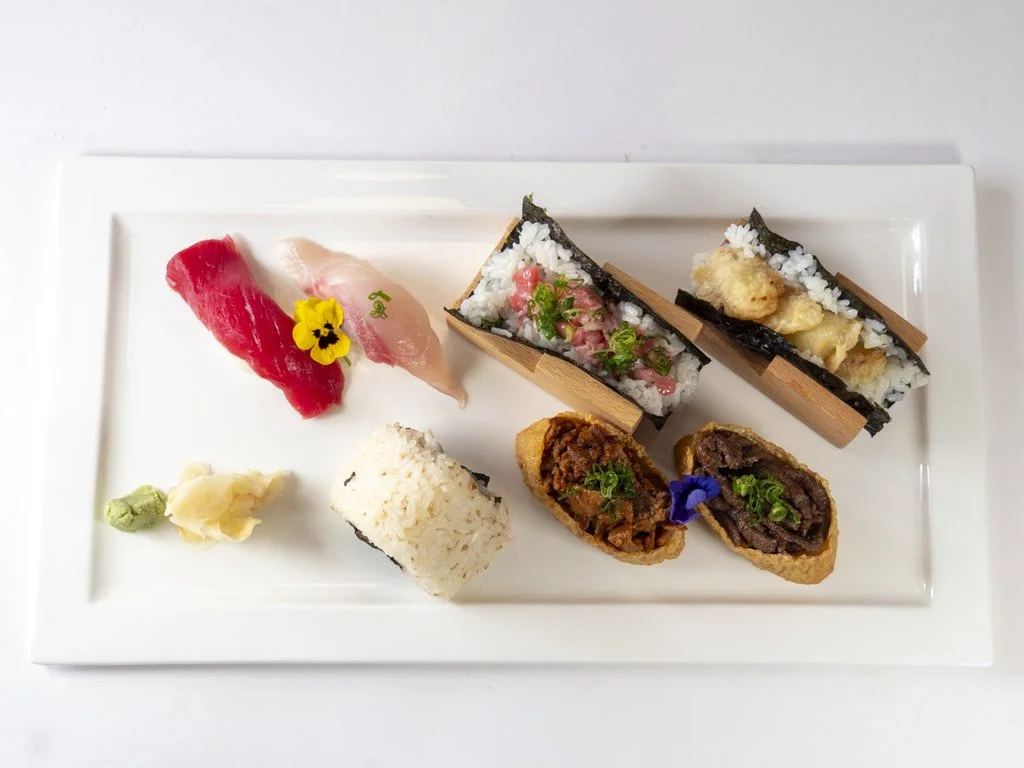 Assorted sushi and sashimi on a rectangular white plate, including two pieces of nigiri with raw fish, two sushi rolls with seaweed and rice, and two deep-fried rolls with fillings, garnished with wasabi, pickled ginger, and decorative flowers.