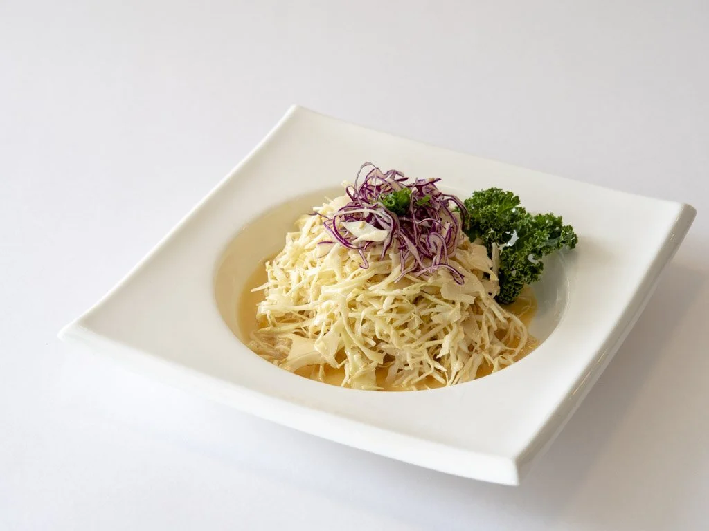 A plate of shredded cabbage salad garnished with purple onion strips, parsley, and a small leaf of green leaf vegetable on a white square dish.