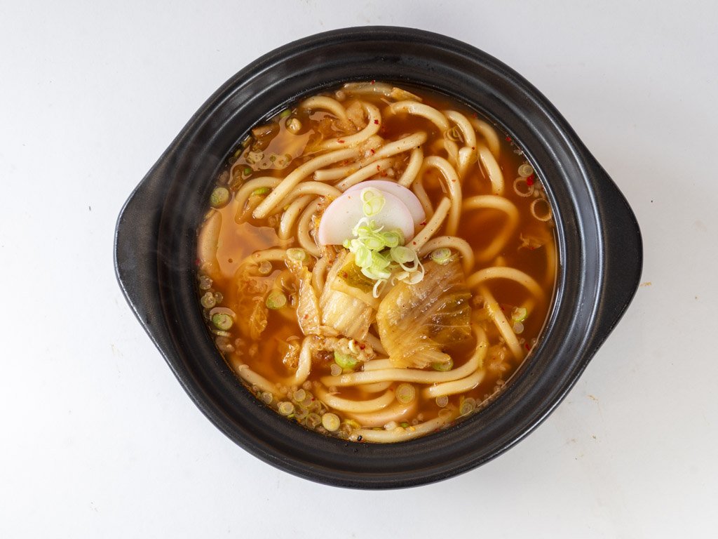 A bowl of Asian noodle soup with sliced vegetables, green onions, and a slice of fish cake, served in a black bowl on a white surface.