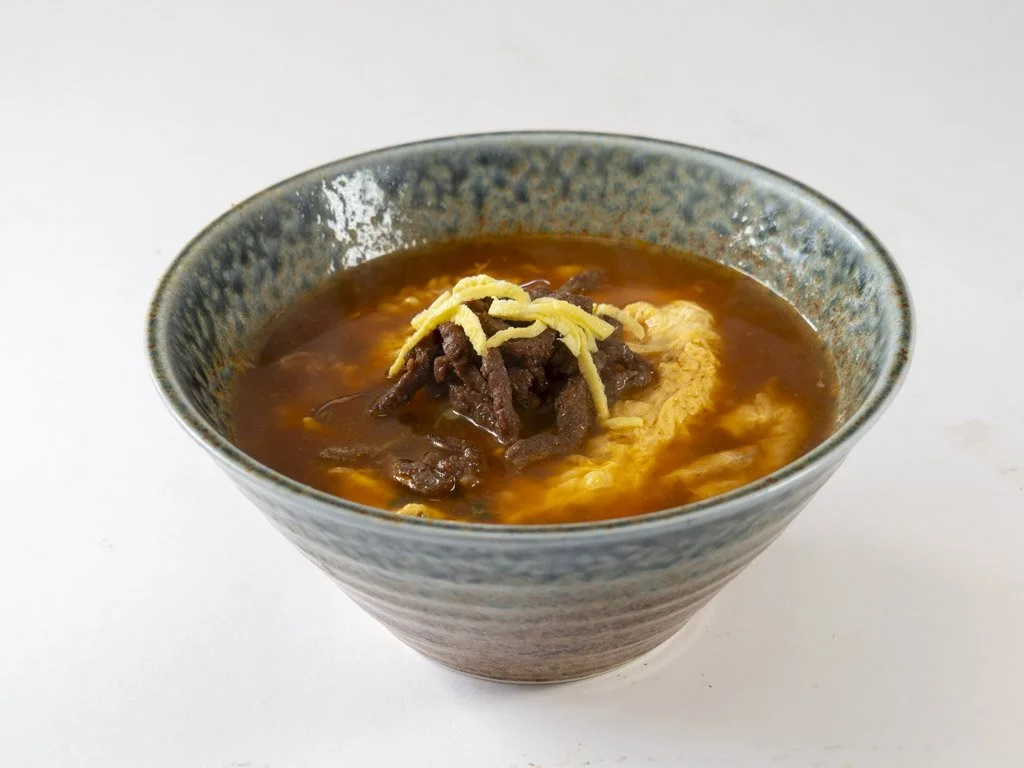 A bowl of hot soup with beef, shredded cheese, and egg.
