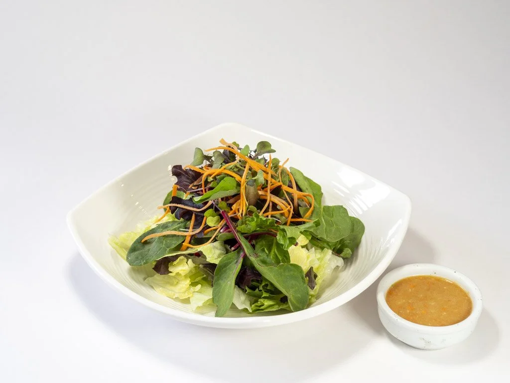 Mixed green salad with shredded carrots served in a white bowl, accompanied by a small bowl of salad dressing.