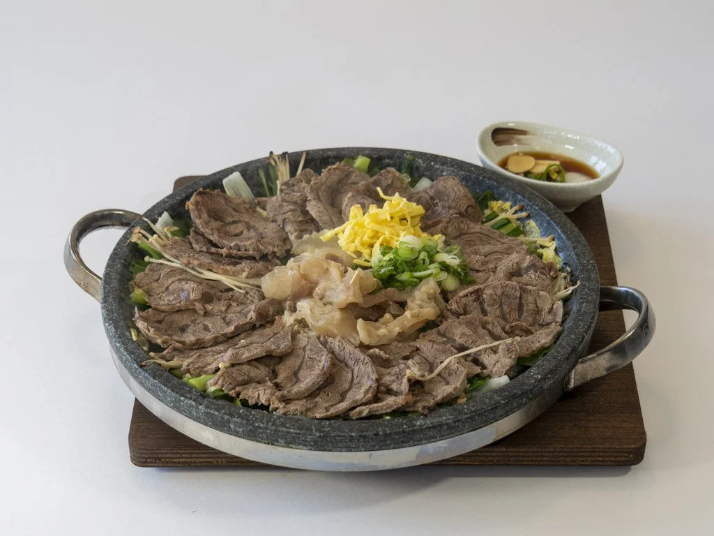 Thinly sliced beef arranged on a hot stone platter with chopped green onions, shredded yellow cheese, and pickled vegetables in the center, served with a small bowl of soy sauce and chili for dipping.