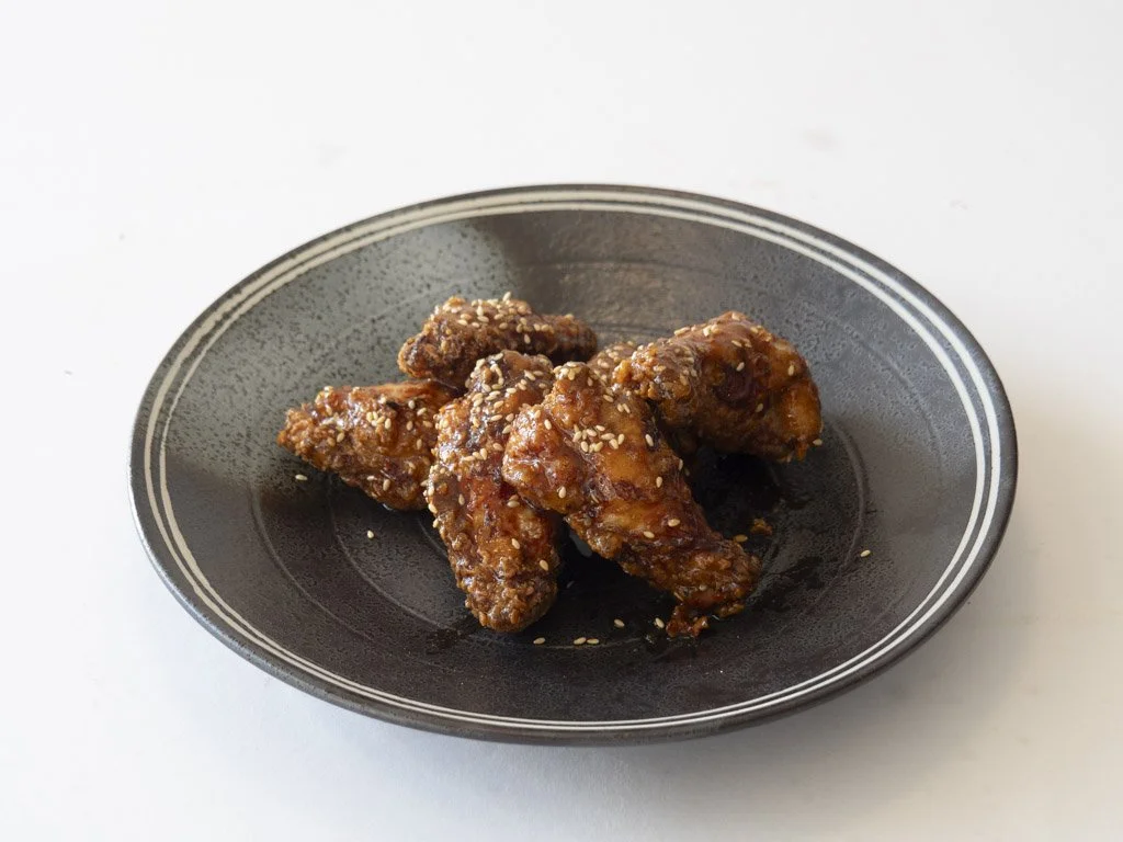 Five pieces of glazed chicken wings sprinkled with sesame seeds on a black oval plate.