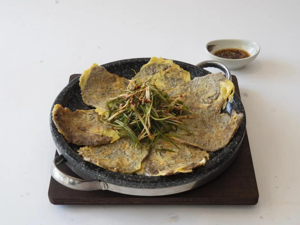 A round black skillet containing sliced pieces of meat topped with green leafy garnish, served on a wooden board with a small bowl of dark dipping sauce beside it.