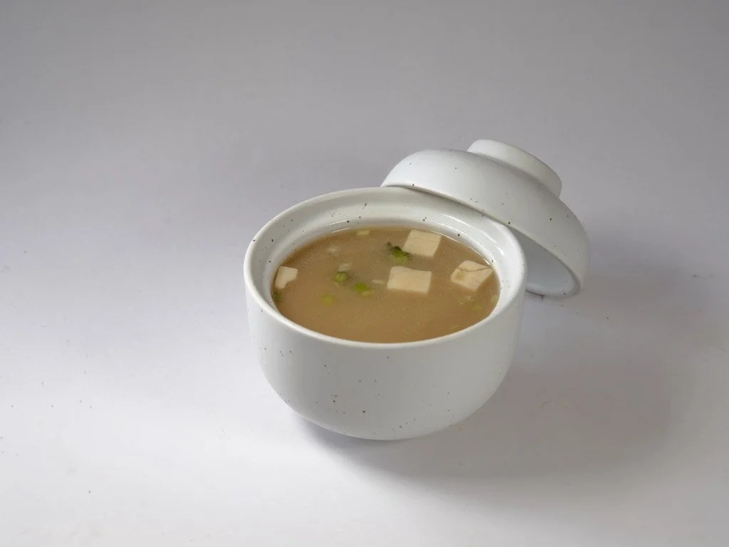 A white ceramic bowl with a lid, filled with soup containing tofu and green onions, placed on a white surface.