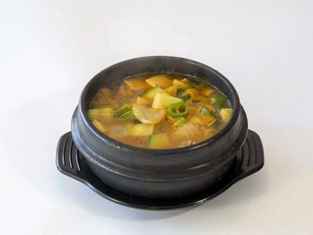A black stone bowl filled with hot soup containing vegetables like zucchini, onions, and green onions on a white surface.