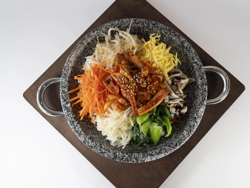 A bowl of bibimbap with rice, carrot, bean sprouts, shredded egg, mushrooms, green vegetables, and seasoned meat garnished with sesame seeds, served on a dark wooden tray.
