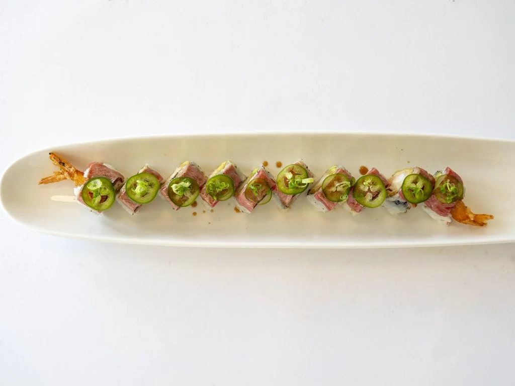 Sushi roll with slices of jalapeño peppers on top, served on a long white plate.