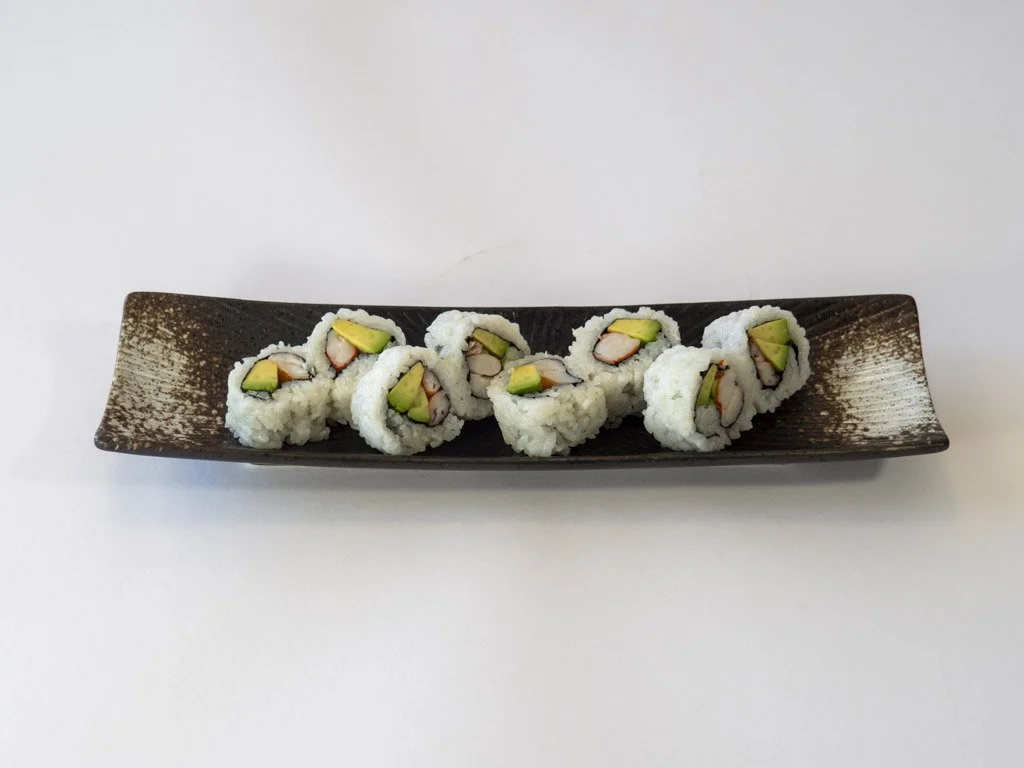 Seven sushi rolls with avocado and crab inside, arranged on a long dark rectangular plate.