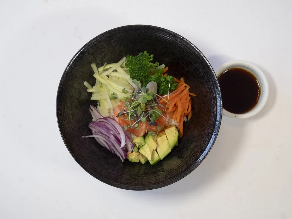 A bowl of assorted fresh vegetables including avocado, red onion, shredded carrot, broccoli, celery, and microgreens, with a small cup of soy sauce on a white surface.