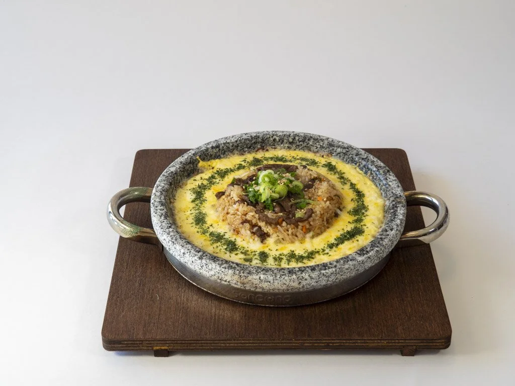 A bowl of rice topped with beef and chopped green onions, served in a stoneware dish with handles on a wooden board.