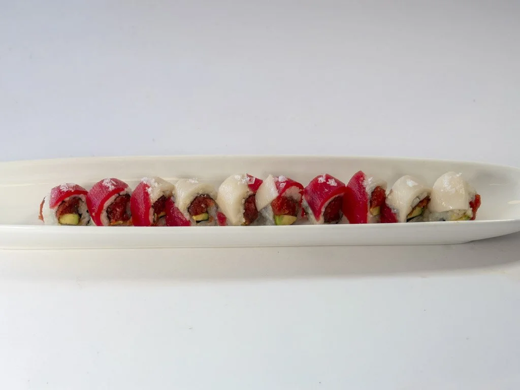 A long white plate with ten sushi rolls, each topped with red and white fish, on a white surface with a plain white background.