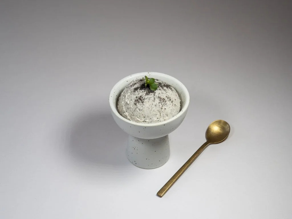 A small white cup of cookies and cream ice cream topped with crumbled cookies and a small green herb, accompanied by a gold spoon, on a plain white surface.