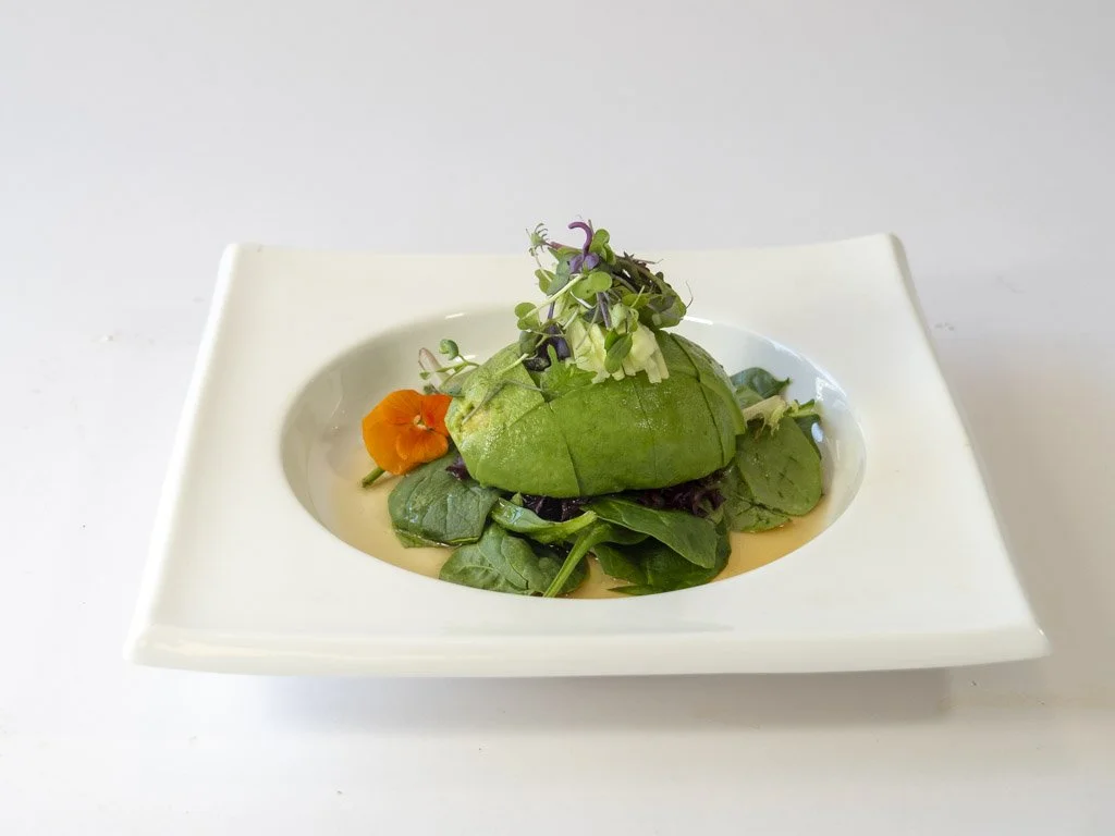 A dish with a green avocado, mixed greens, and edible flowers on a white square plate.