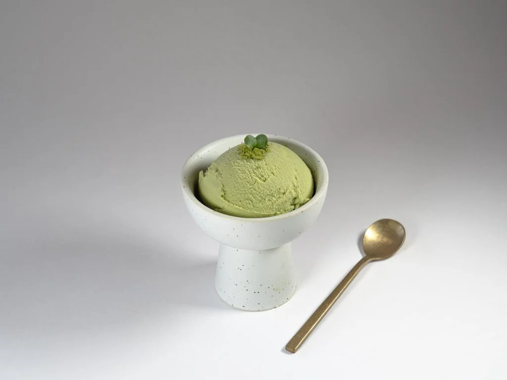 A scoop of green matcha ice cream in a white cup with a small green garnish on top, a gold spoon beside it, on a plain gray background.