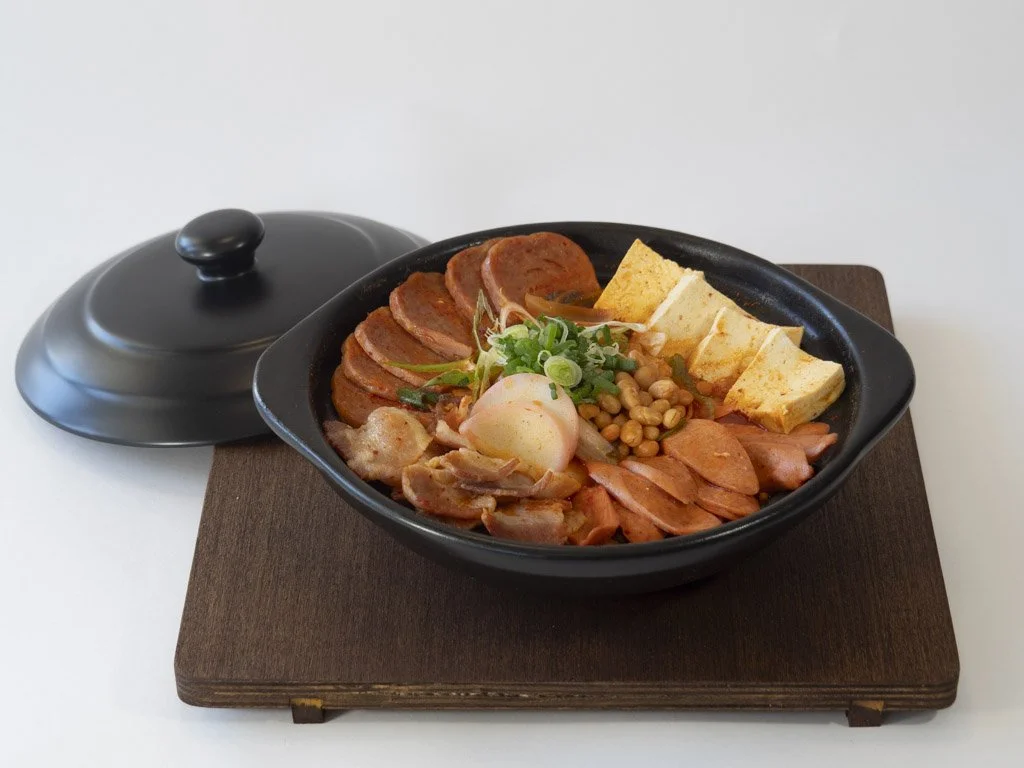 A black bowl of assorted Japanese-style foods on a wooden tray, with sliced meats, tamagoyaki, vegetables, and garnishes.