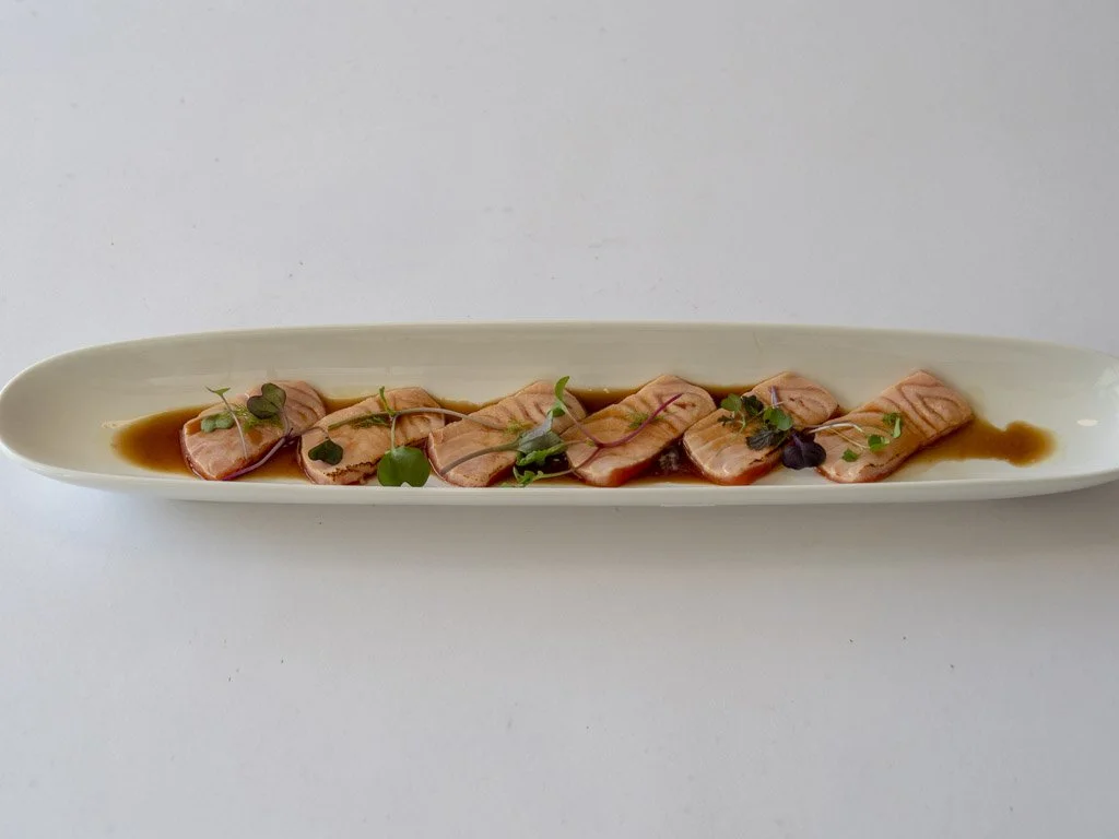 Slices of raw salmon sashimi garnished with microgreens and drizzled with soy sauce on an elongated white platter.