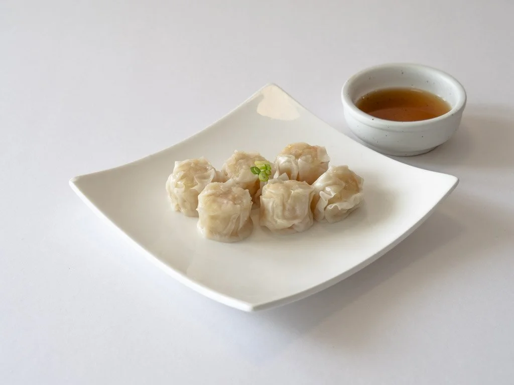 Plate with six shumai dumplings and a small bowl of dipping sauce on a white background.